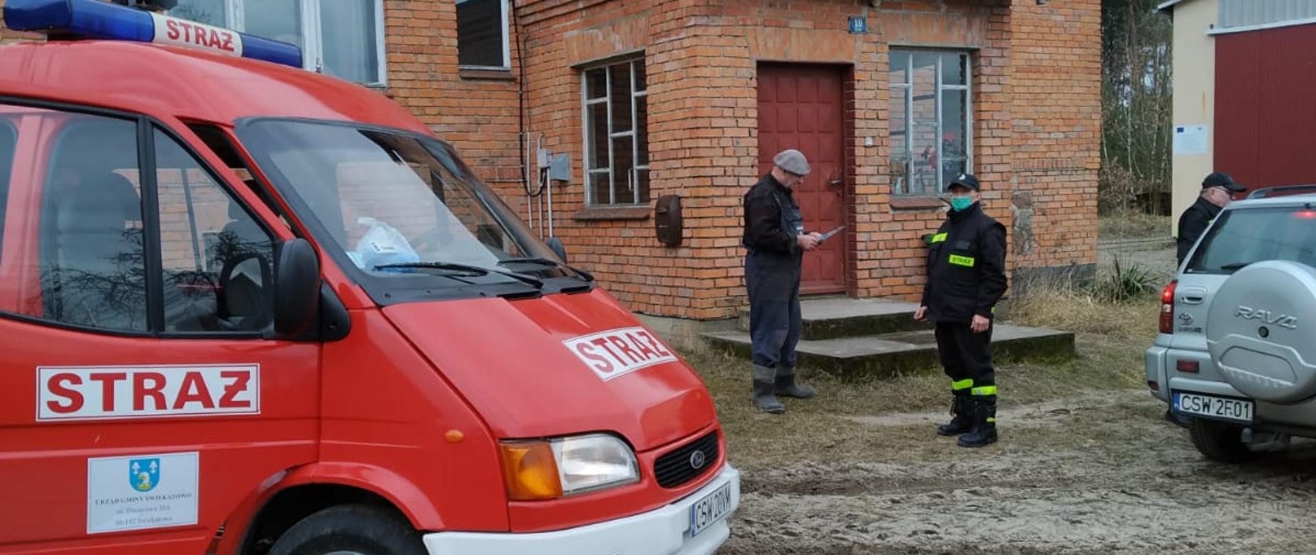 Po lewej stronie widać przód samochodu pożarniczego Ford Transit Ochotniczej Straży Pożarnej w Szewnie stojący na posesji mieszkańca miejscowości w tlen widać dom jednorodzinny zbudowany z cegły. Przed domem stoi strażak Ochotniczej Straży Pożarnej w Szewnie a przed nim właściciel posesji czytający ulotkę. Po prawej widać tył samochodu osobowego przy którym stoi mężczyzna. 