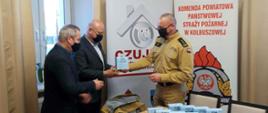 Na zdjęciu 3 mężczyzn: strażak w umundurowaniu w kolorze piaskowymi oraz dwóch w garniturach. Jednemu z mężczyzn strażak przekazuje jedną z czujek tlenku węgla. Na stole pozostałe przekazywane urządzenia oraz inne materiały. Zdjęcie w pomieszczeniu z dużym oknem i długimi zasłonami. W rogu banery z napisami: Komenda Powiatowa Państwowej Straży Pożarnej w Kolbuszowej wraz z logo PSP oraz Czujka na straży Twojego bezpieczeństwa.