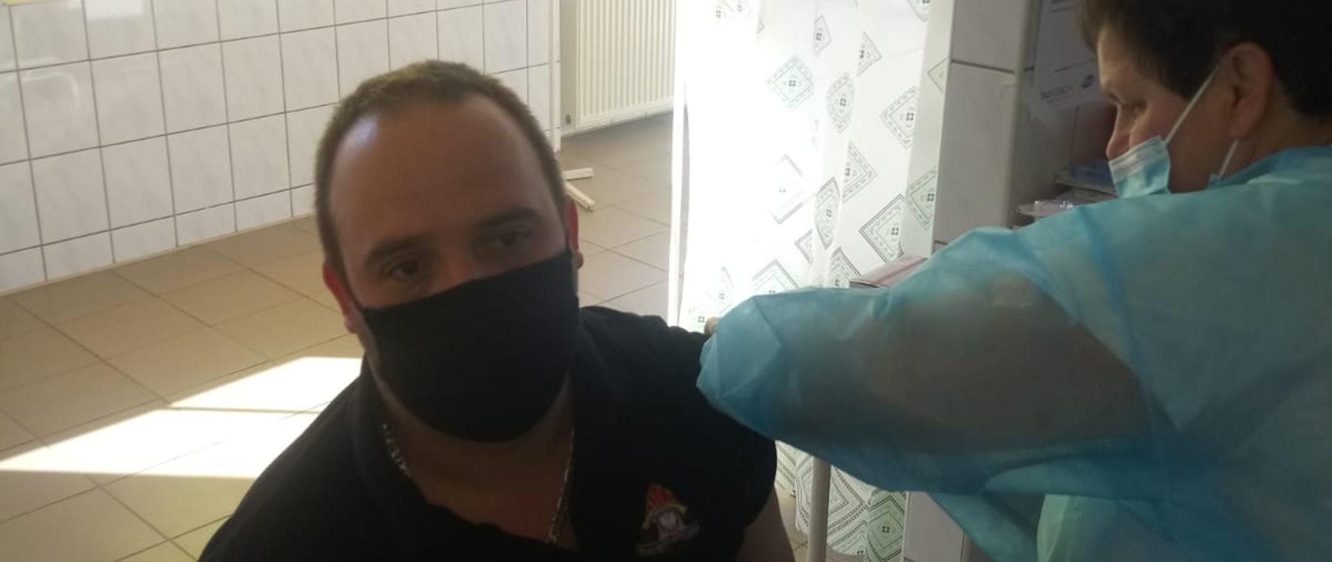 Zdjęcie przedstawia strażaka który jest szczepiony przeciwko covid dziewiętnaście jednej z wielu placówek medycznych.