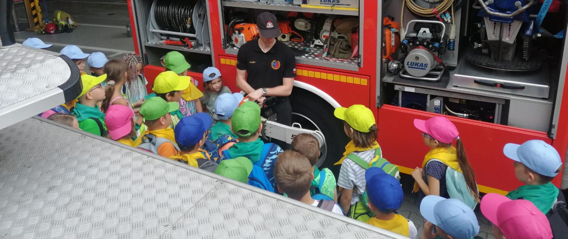 Zdjęcie przedstawi młodzież oglądającą sprzęt pożarniczy. W tle stoi samochód z otwartymi skrytkami na ze sprzętem.