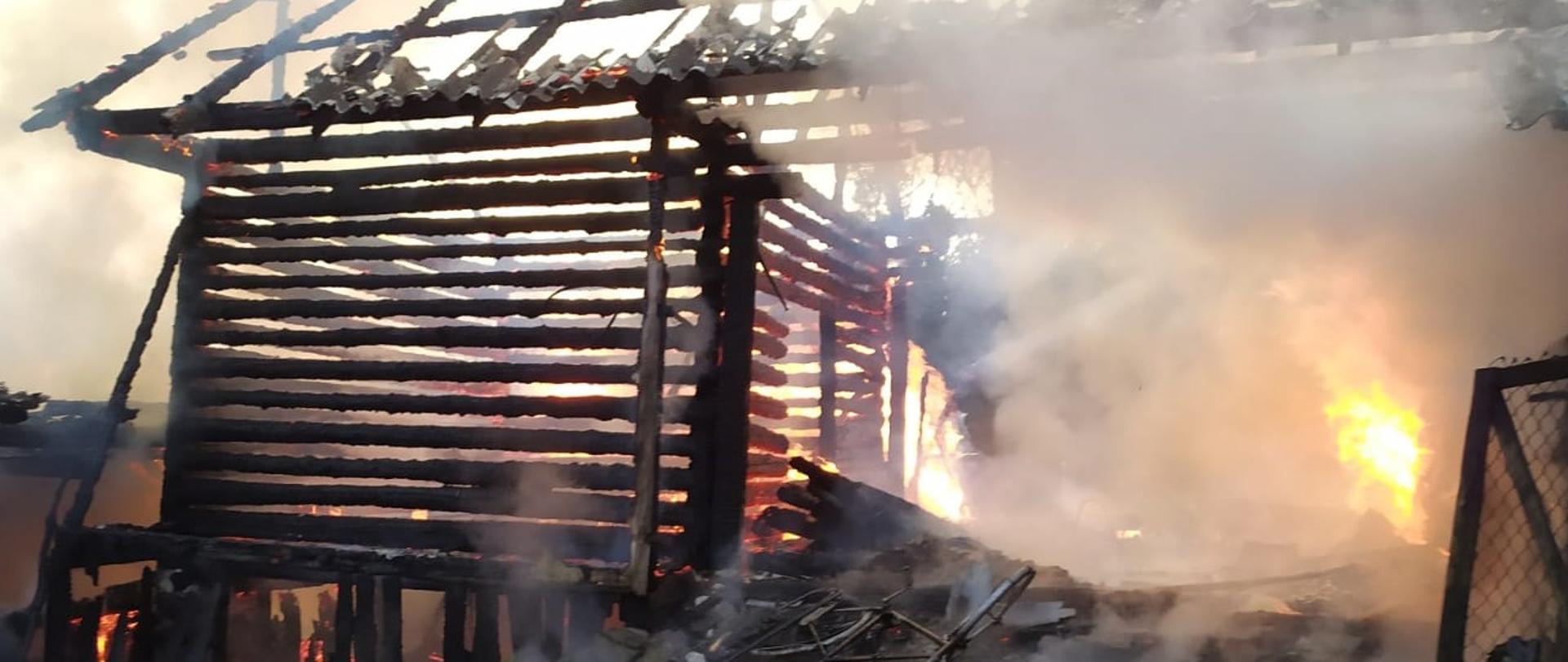Zdjęcie przedstawia konstrukcję drewniana budynku gospodarczego, która objęta jest częściowo płomieniami. Pokrycie dachu z płyt eternitu zostało zniszczone całkowicie. 
