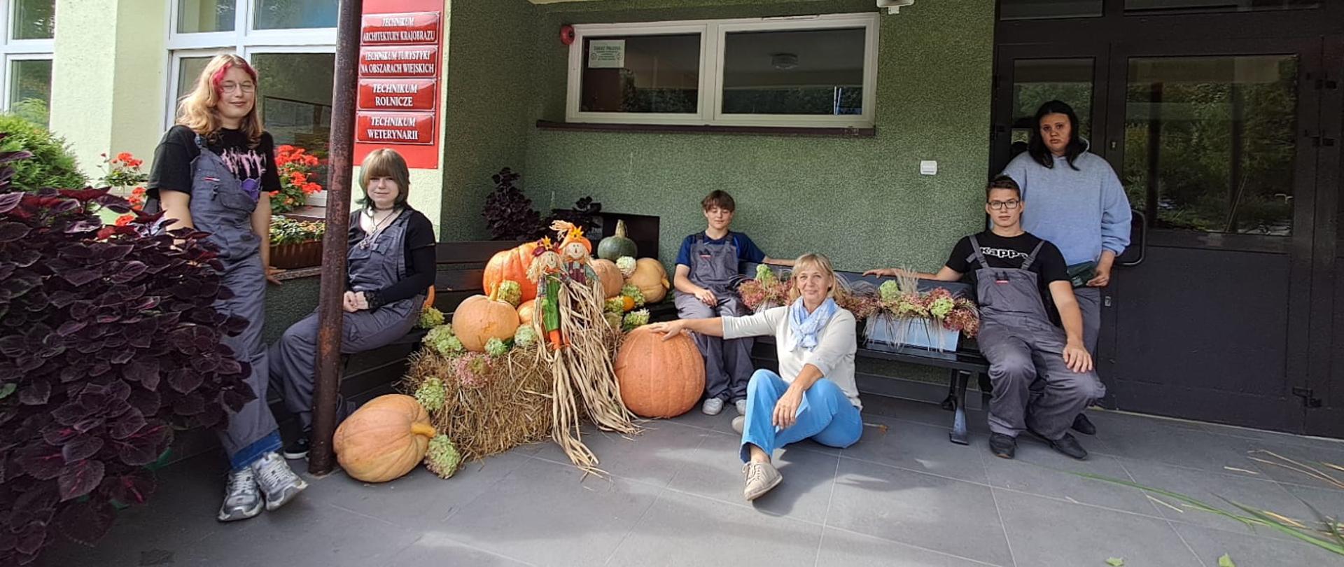 Wspólne pamiątkowe zdjęcie grupy uczniów pozujących przy bogatej dekoracji z dyń, słomy i kwiatów przed wejściem do budynku.