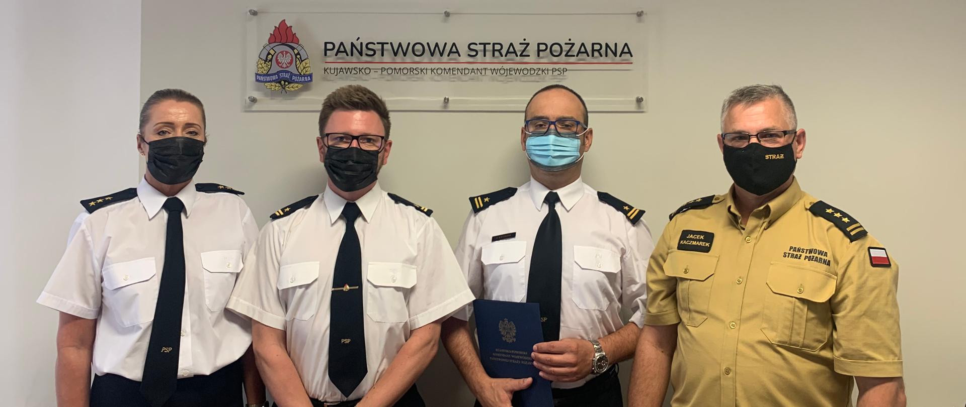 Na zdjęciu czworo funkcjonariuszy ustawionych w szeregu, jeden z nich trzyma teczkę. Nad funkcjonariuszami na ścianie znajduje się logo i napis Państwowa Straż Pożarna, godło Polski oraz herb województwa kujawsko-pomorskiego. Zdjęcie wykonano w gabinecie komendanta.