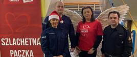 Zdjęcie wykonane na terenie jednej ze szkół biorących udział w ramach akcji szlachetna paczka. Na zdjęciu widać klatkę schodową wewnątrz obiektu. Przy schodach stoją od lewej policjanci, wolontariuszka szlachetnej paczki oraz strażak państwowej straży pożarnej. Funkcjonariusze wspierają wolontariuszy w ramach akcji przekazywania darów osobom potrzebującym.
