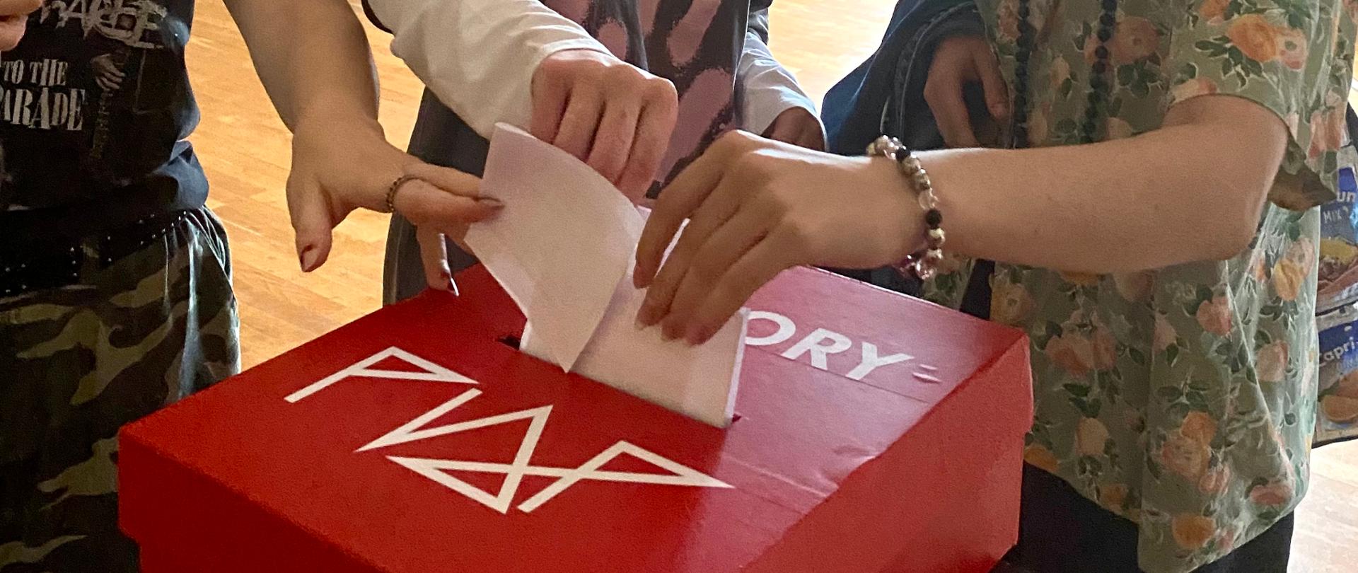 Trzy uczennice wrzucające jednocześnie swoje karty wyborcze do czerwonej urny wyborczej z białym logo szkoły i napisem wybory. Osoby są uśmiechnięte z wyciągniętymi rękoma w kierunku urny, ubrane we wzorzyste koszulki.