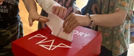 Czerwona urna wyborcza z białym logo szkoły i napisem wybory z dłońmi które wrzucają głosy do otworu w urnie.