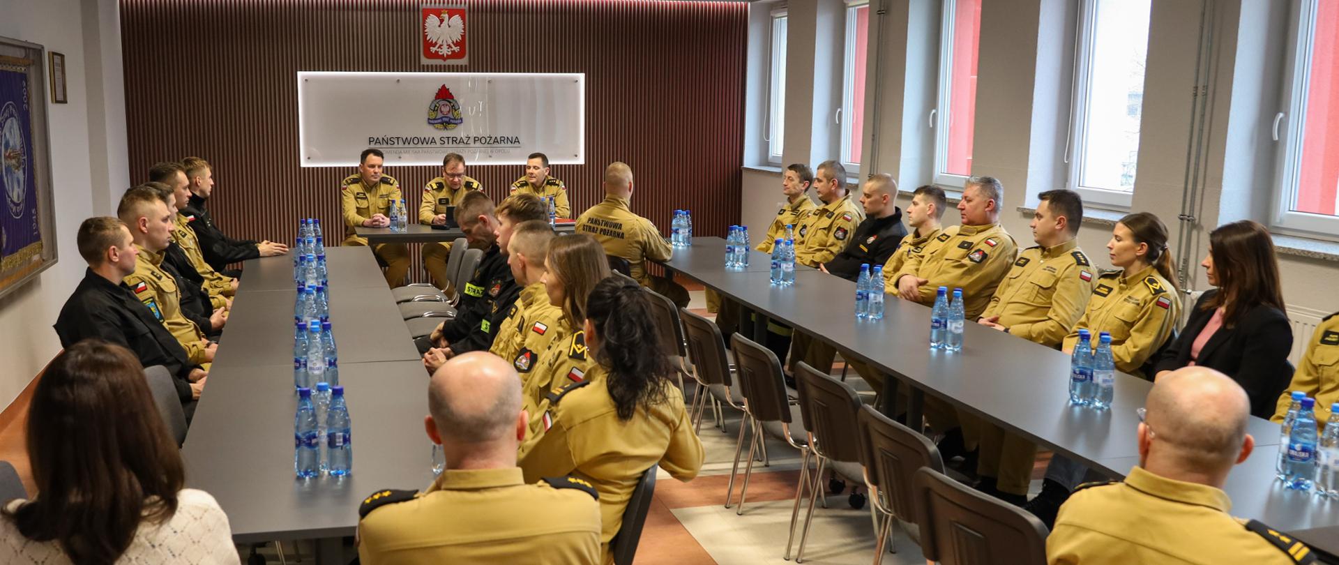 Opolski Komendant Wojewódzki, Zastępca Opolskiego Komendanta Wojewódzkiego oraz Zastępca Komendanta Miejskiego PSP w Opolu oraz kadra KM PSP w Opolu podczas spotkania. W tle godło Polski, logo PSP i napis "PAŃSTWOWA STRAŻ POŻARNA".
