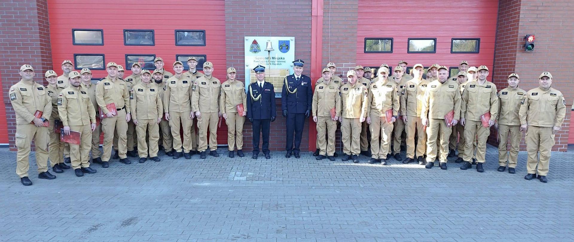 Na zdjęciu przedstawiono grupę funkcjonariuszy Państwowej Straży Pożarnej podczas zbiórki przed siedzibą Komendy Miejskiej PSP. Strażacy w umundurowaniu służbowym stoją w zwartym szyku, a w centralnej części widoczni są przełożeni w umundurowaniu wyjściowym.
Część funkcjonariuszy trzyma dokumenty lub materiały szkoleniowe, co wskazuje na zakończenie szkolenia, odprawę lub uroczyste wręczenie zaświadczeń. W tle widoczne są bramy garażowe jednostki oraz tablica identyfikacyjna komendy.