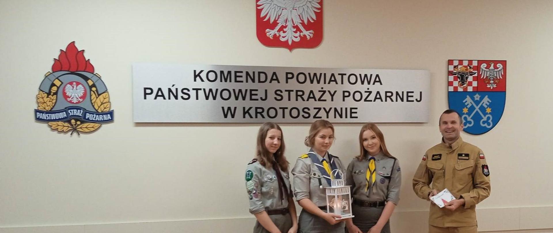 Zdjęcie przedstawia trzy harcerki oraz strażaka w mundurze koloru piaskowego. Osoby stoją na tle ściany z napisem Komenda Powiatowa Państwowej Straży Pożarnej w Krotoszynie, godła Polski oraz loga PSP i Starostwa Powiatowego podczas uroczystego przekazywania lampionu z Betlejemskim Światełkiem Pokoju.