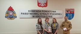 Zdjęcie przedstawia trzy harcerki oraz strażaka w mundurze koloru piaskowego. Osoby stoją na tle ściany z napisem Komenda Powiatowa Państwowej Straży Pożarnej w Krotoszynie, godła Polski oraz loga PSP i Starostwa Powiatowego podczas uroczystego przekazywania lampionu z Betlejemskim Światełkiem Pokoju.