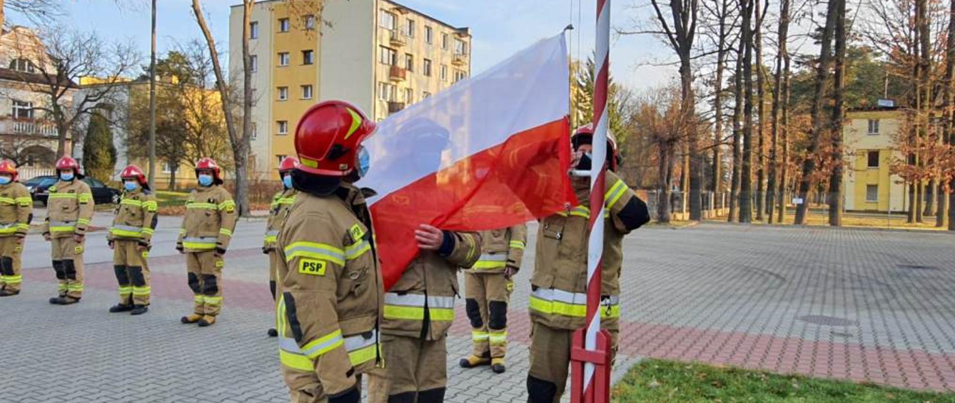 Zdjęcie przedstawia trzech funkcjonariuszy Państwowej Straży Pożarnej. Funkcjonariusze ubrani są w ubrania specjalne w kolorze żółtym. Na głowach mają czerwone hełmy. Dwóch z nich przygotowuje flagę do wciągnięcia na maszt. Zdjęcie wykonane przed Jednostką Ratowniczo – Gaśniczą numer 4 w Pionkach. 