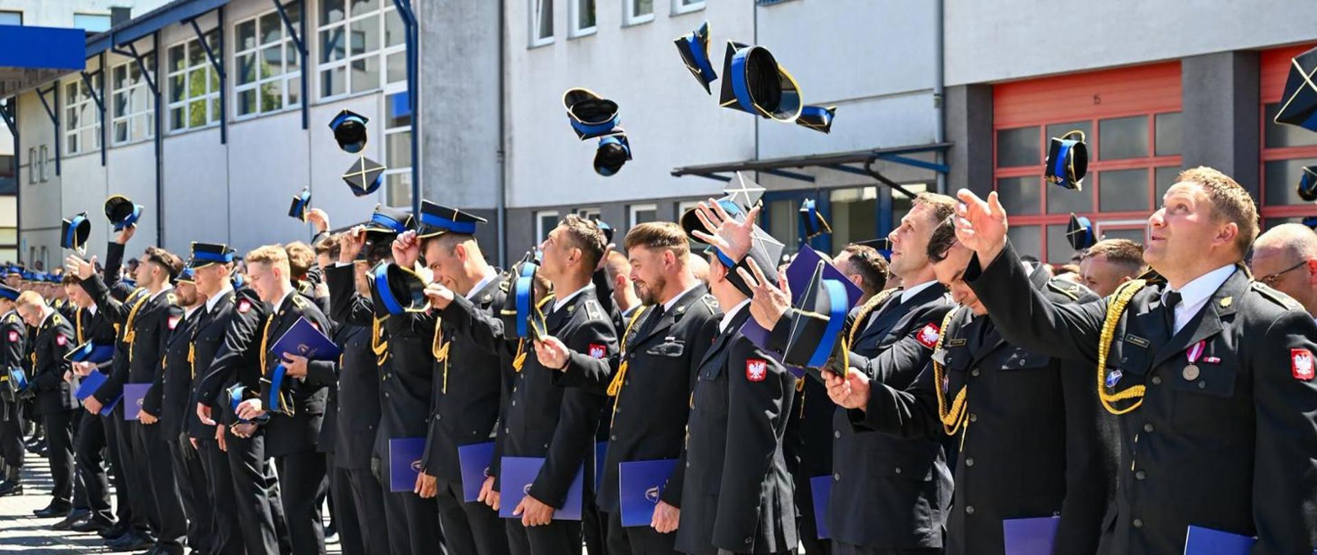 Zdjęcie przedstawia uroczystość ukończenia szkoły przez grupę młodych strażaków. Na pierwszym planie widać szereg mężczyzn w galowych mundurach Państwowej Straży Pożarnej – ciemnogranatowych kurtkach z czerwonymi wypustkami, złotymi guzikami i sznurami galowymi oraz białych orzełkach na czapkach i emblematach na rękawach. Strażacy stoją w rzędach na placu apelowym przed budynkiem komendy, w tle widać betonowe ściany i czerwone bramy garażowe. Niektórzy z nich z radością podrzucają w górę czapki rogatywki, inne czapki są już w locie, tworząc dynamiczną kompozycję na tle nieba. Większość absolwentów uśmiecha się szeroko i patrzy w górę za swoimi nakryciami głowy. W dłoniach trzymają niebieskie dyplomy. Zdjęcie jest pełne energii, radości i dumy, upamiętniając ważny moment w życiu młodych strażaków.