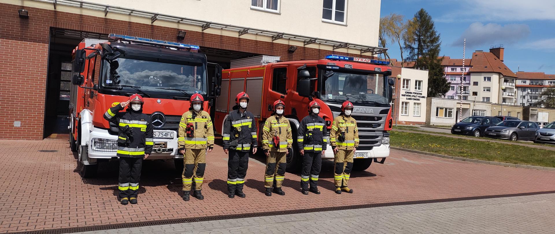 Zdjęcie przedstawia strażaków stojących w szeregu przed garażem straży pożarnej. Za nimi stoją wozy strażackie.