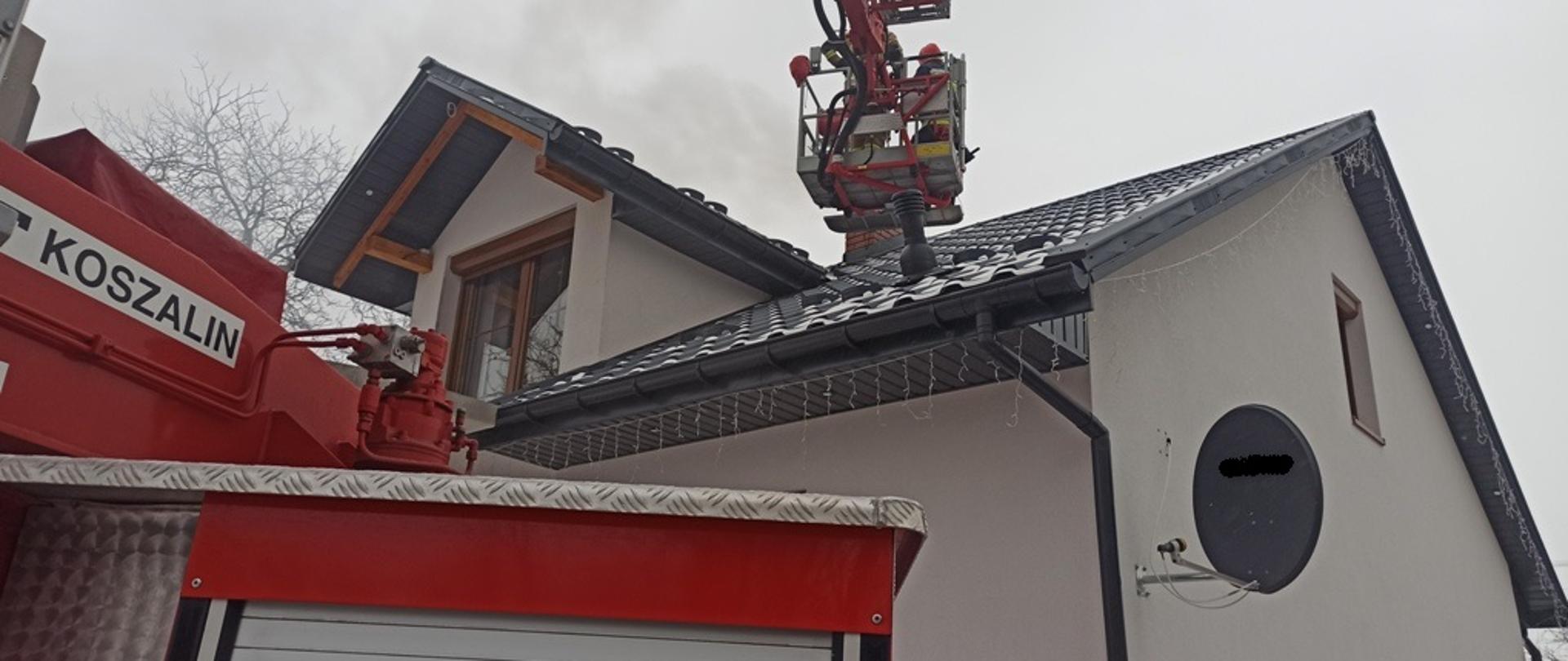 Na zdjęciu pojazd pożarniczy specjalny-podnośnik, w koszu podnośnika strażacy prowadzący działania gaśnicze przewodu kominowego, w tle zdjęcia budynek mieszkalny.