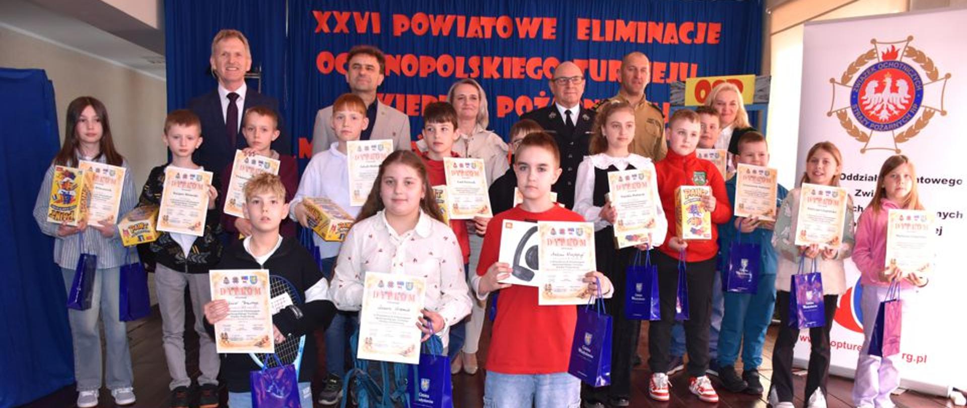 Grupa dzieci stoi na scenie z dyplomami i nagrodami, pozując do zdjęcia z organizatorami po zakończonym konkursie. 