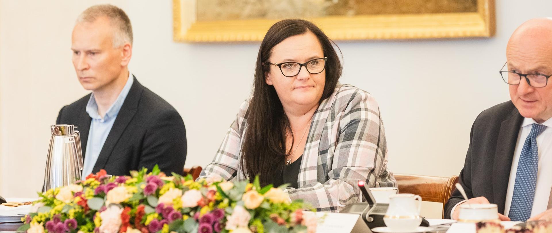 Wiceminister Małgorzata Jarosińska-Jedynak siedzi przy stole z kwiatami. Obok niej dwóch mężczyzna.