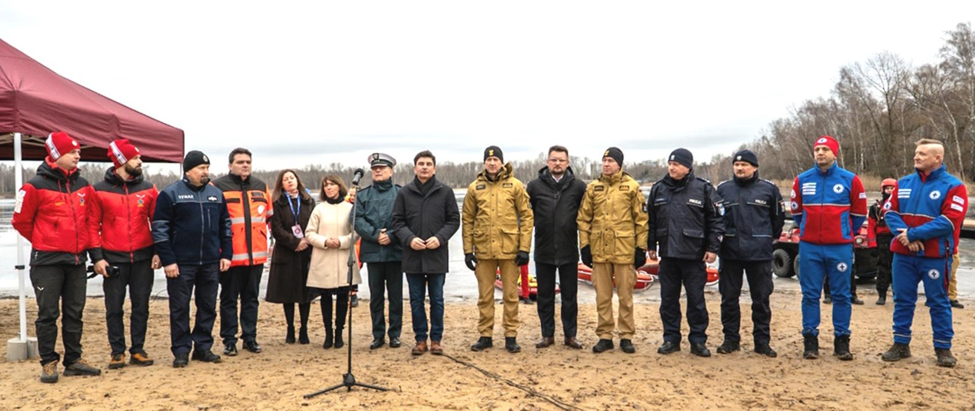 Grupa kilkunastu osób stojących w jednym rzędzie na piaszczystym brzegu zbiornika wodnego.
Osoby są ubrane głównie w stroje służbowe i ratownicze — część ma czerwone kurtki, inni granatowe lub brązowe mundury, a kilka osób cywilne płaszcze. W centrum ustawione są mikrofony na statywach.
Po lewej stronie stoi czerwony namiot, a po prawej widać sprzęt ratowniczy i pojazd terenowy. 