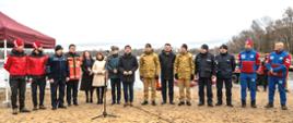 Grupa kilkunastu osób stojących w jednym rzędzie na piaszczystym brzegu zbiornika wodnego.
Osoby są ubrane głównie w stroje służbowe i ratownicze — część ma czerwone kurtki, inni granatowe lub brązowe mundury, a kilka osób cywilne płaszcze. W centrum ustawione są mikrofony na statywach.
Po lewej stronie stoi czerwony namiot, a po prawej widać sprzęt ratowniczy i pojazd terenowy. 