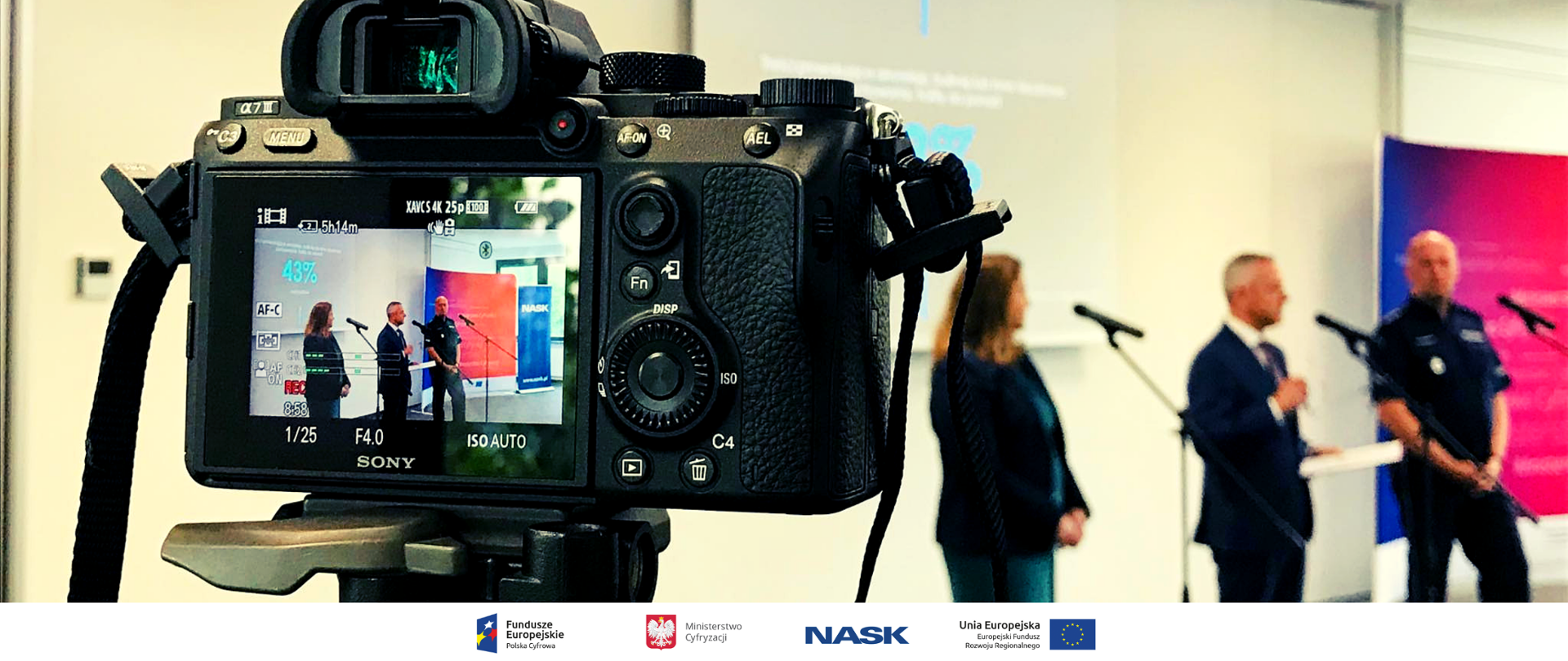 Konferencja prasowa. Aparat fotograficzny skierowany na trzy osoby. 