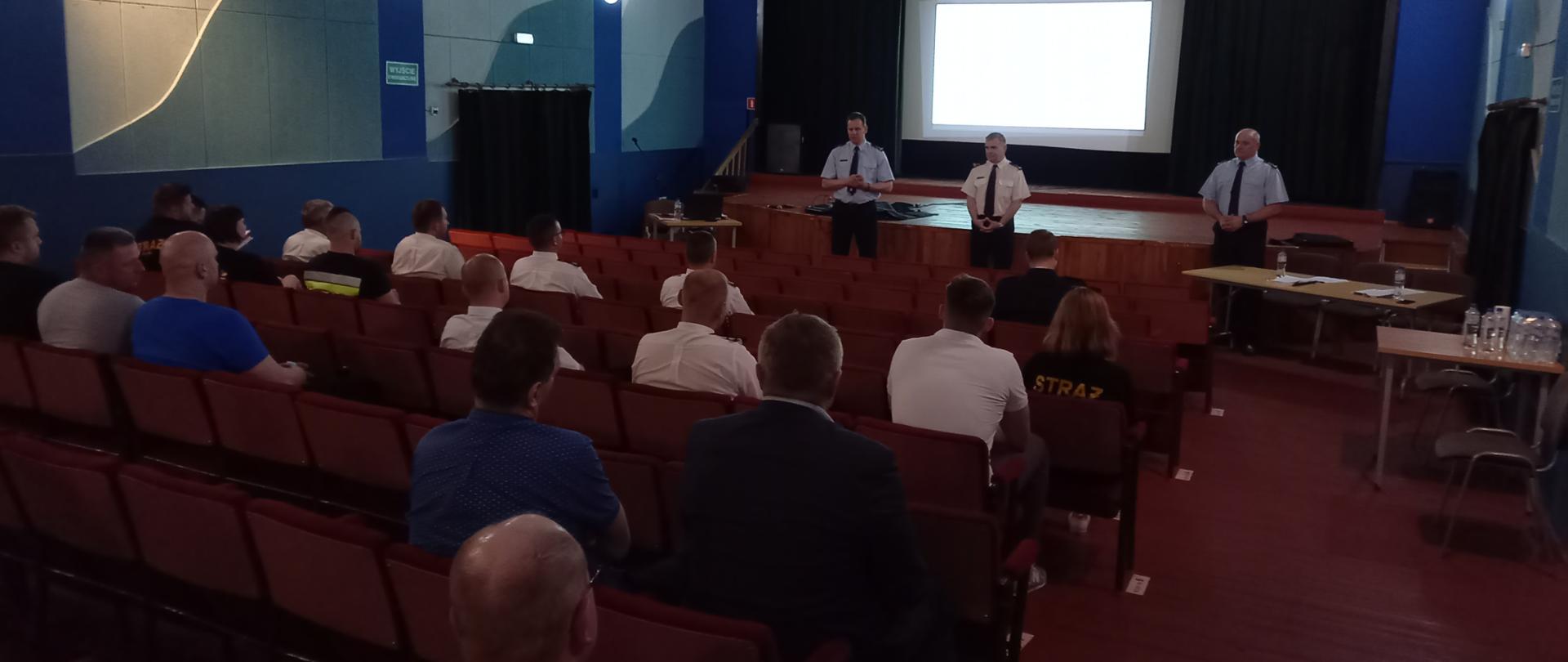 Widok wnętrza sali kinowej. Na fotelach siedzący przedstawiciele OSP. Z przodu trzech funkcjonariuszy PSP - Lubuski Komendant Wojewódzki PSP wraz z zastępcą oraz Komendant Powiatowy PSP w Słubicach, zwróceni twarzą do siedzących druhów.