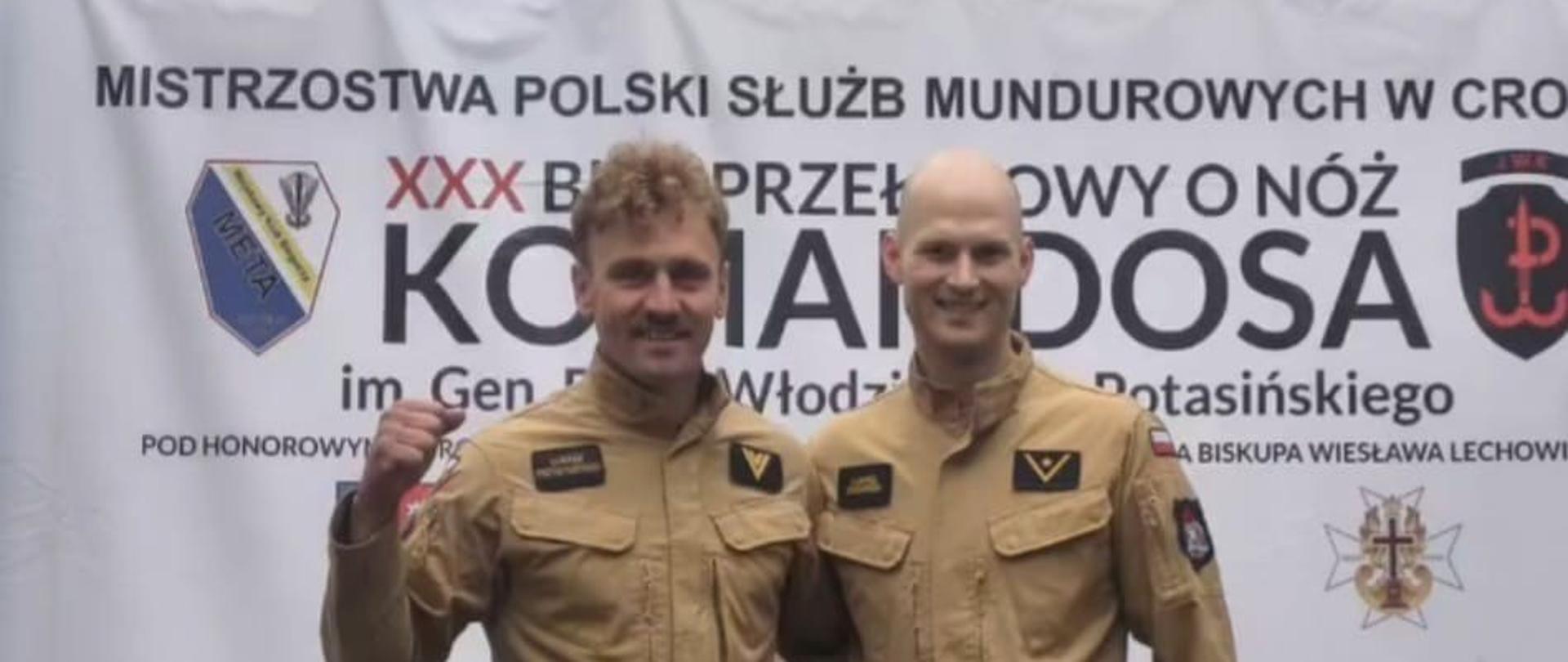 Funkcjonariusze KP PSP w Mławie, Łukasz Przybyszewski i Łukasz Zdziarski, pozują do zdjęcia na tle baneru XXX Biegu Przełajowego „O Nóż Komandosa”. Jeden ze strażaków unosi kciuk w geście zwycięstwa.