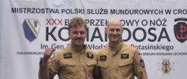 Funkcjonariusze KP PSP w Mławie, Łukasz Przybyszewski i Łukasz Zdziarski, pozują do zdjęcia na tle baneru XXX Biegu Przełajowego „O Nóż Komandosa”. Jeden ze strażaków unosi kciuk w geście zwycięstwa.