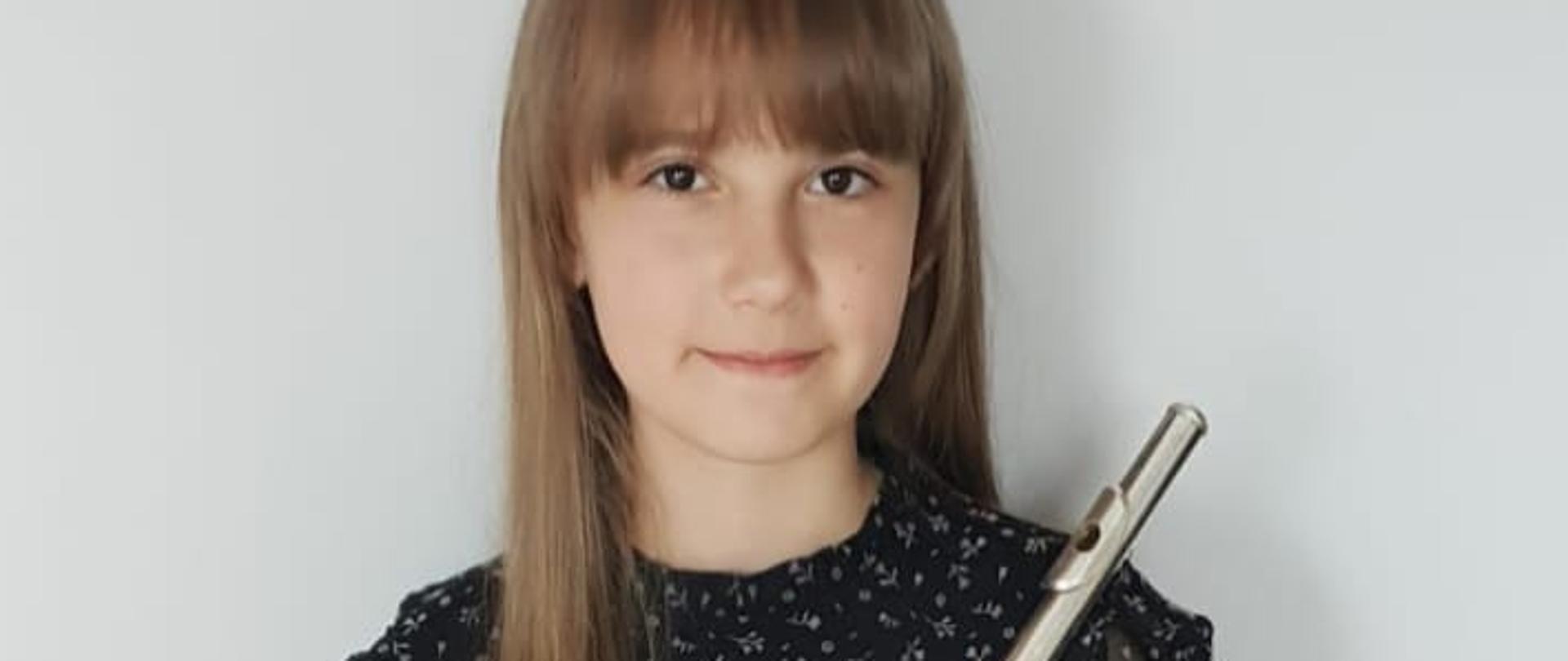 zdjęcie przedstawia dziewięcioletniom dziewczynę o brązowych włosach w czarnej sukience w kwiaty, trzymającą w obu rękach flet na jasnym tle