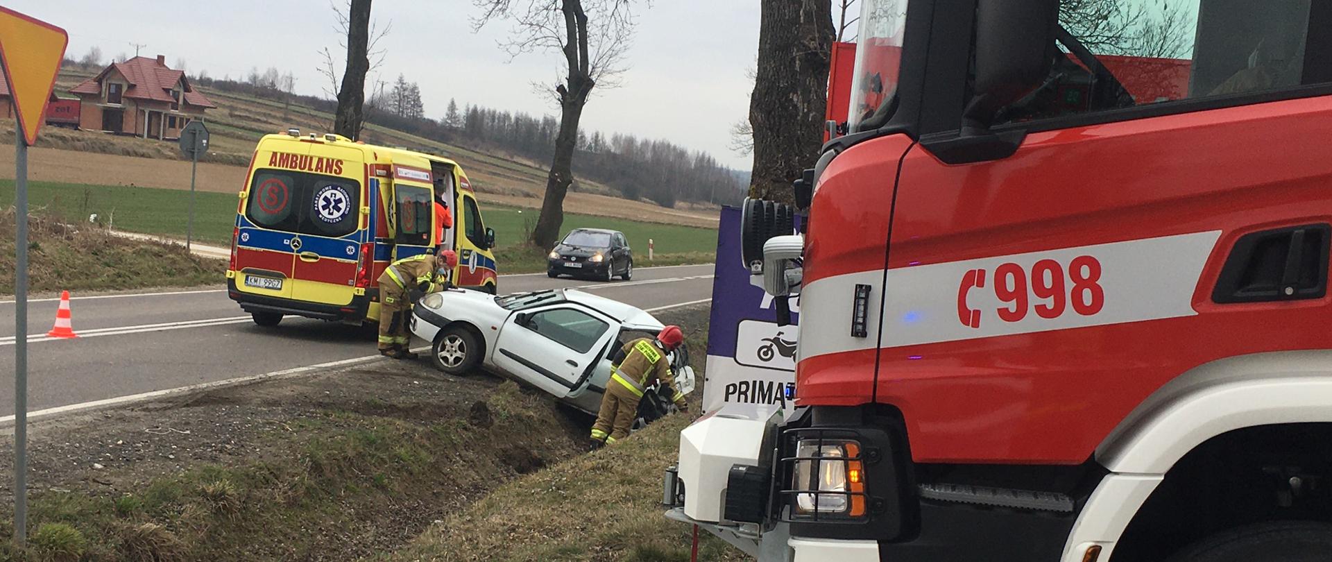na miejscu widoczne jest jeden samochód PSP, na pasie jezdni karetka Zespołu Ratownictwa Medycznego w której ratownicy medyczni zabezpieczaj osobę poszkodowaną oraz samochód osobowy ,który leży w rowie z widocznymi uszkodzeniami karoserii przy nim dwóch strażaków PSP