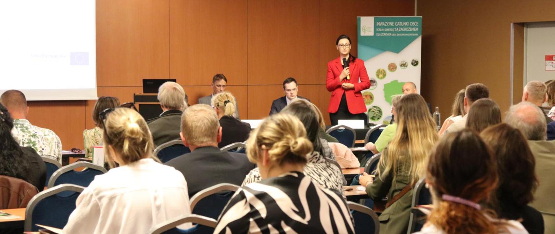 Sala konferencyjna. Trzy osoby przy stoliku twarzami zwrócone do uczestników konferencji. Jedna z z osób za stołem stoi i przemawia do mikrofonu. W tle plansze z napisami.
