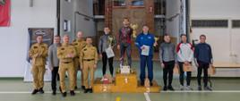W rzędzie stoją funkcjonariusze Państwowej Straży Pożarnej, oraz zaproszenie oficjele w środku na podium stoją sportowcy w dresach w rękach trzymają dyplomy i puchary. Z tyłu widać wspinalnię.