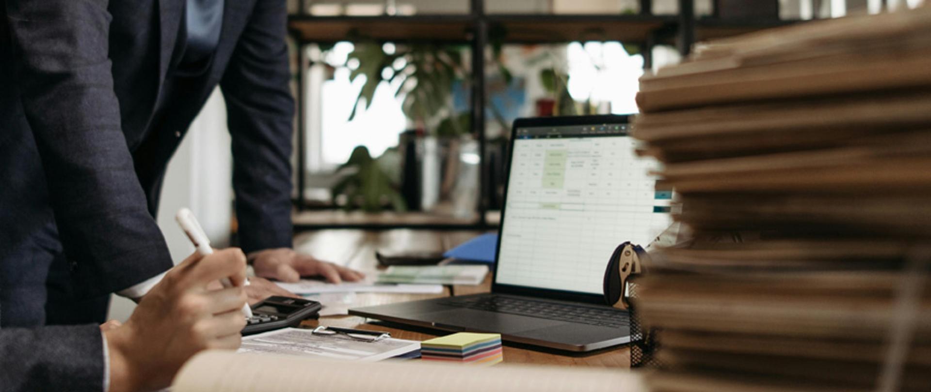 Biurko z laptopem, dokumentami i kalkulatorem, przy którym pracują osoby analizujące dane w arkuszu kalkulacyjnym.