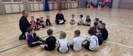 Pogadanka młodych zawodników ze strażakiem na temat zagrożeń mogące wystąpić zarówno w domu, jak i w codziennym życiu. Dzieci siedzą na parkiecie hali sportowej w kółku wysłuchując porad strażaka.