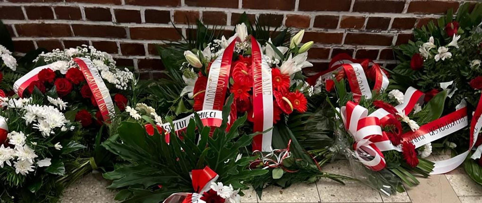 wieńce biało czerwoone z biało czerwonymi wstęgami leżąc pod ścianą pamięci