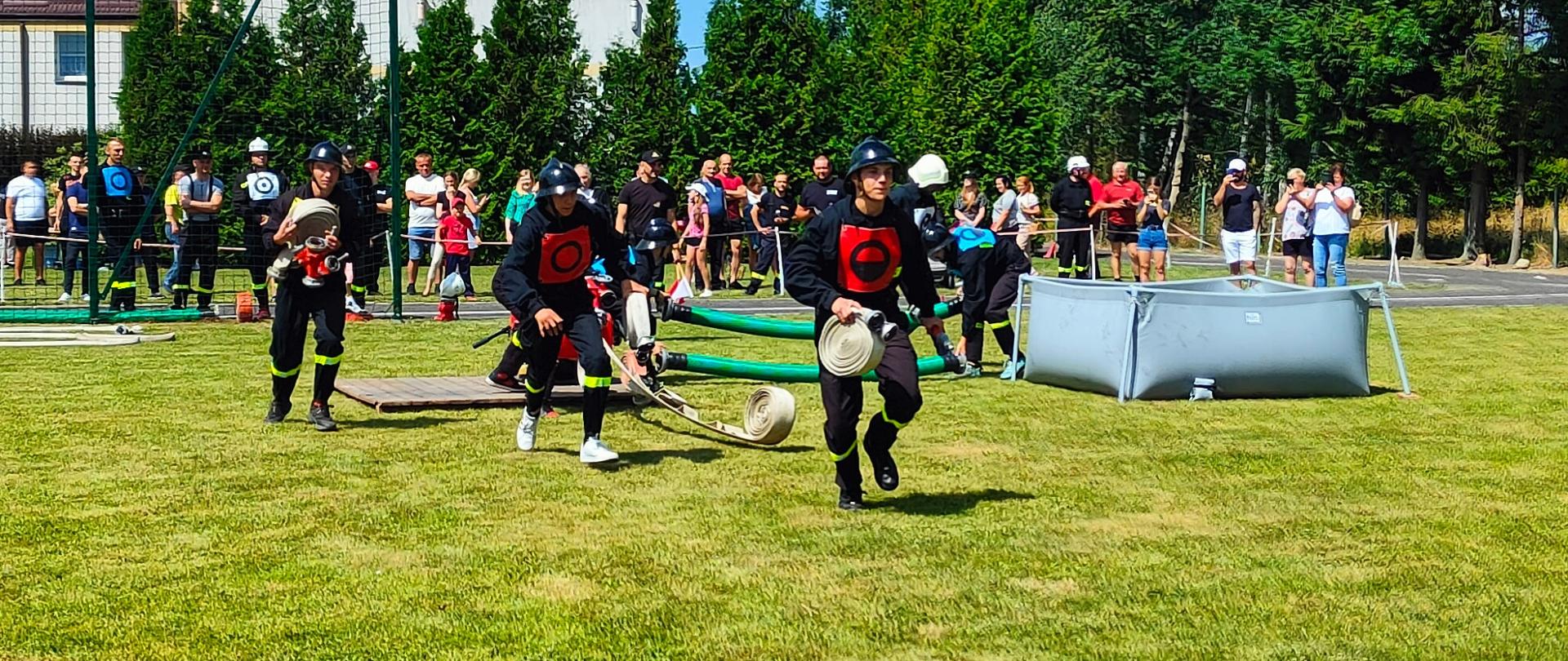 Gminne Zawody Sportowo – Pożarnicze jednostek OSP z terenu gminy Repki 2023 - na zdjęciu strażacy na zawodach na boisku trawiastym biegną z wężami