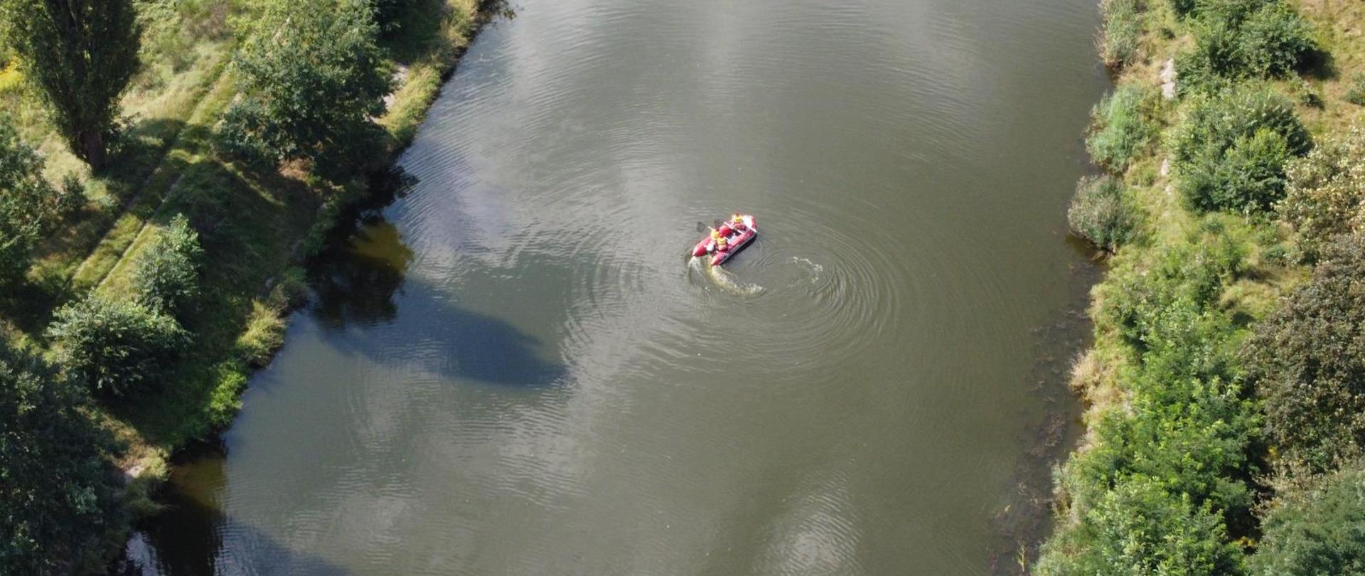 Zdjęcie przedstawia fragment rzeki. Na rzece widać płynący ponton wodny, w którym są trzy osoby