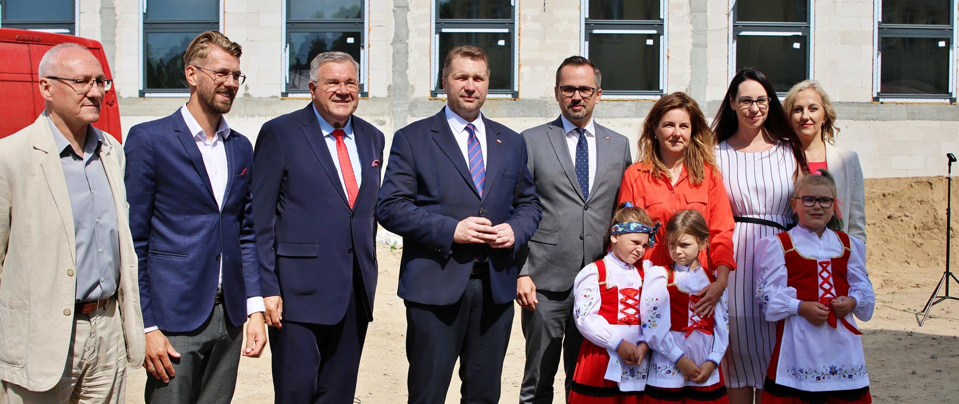 W ostrym słońcu na dworze stoi grupa ludzi, przed nimi trzy małe dziewczynki w biało-czerwonych strojach, w tle widać budynek w trakcie budowy.