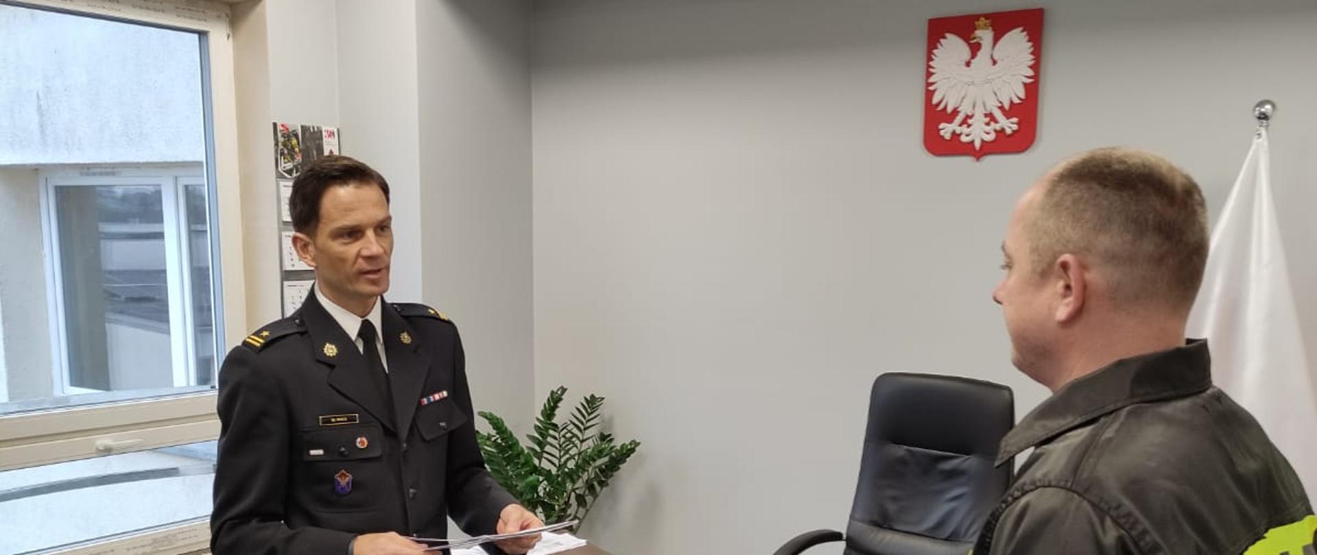 Komendant Powiatowy PSP w umundurowaniu wyjściowym składa gratulacje funkcjonariuszowi PSP ubranemu w czarne umundurowanie koszarowe. Panowie stoją w gabinecie, obok biurko. Na Ścianie w tle godło RP.