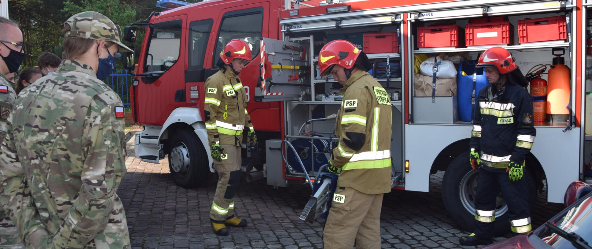 Na zdjęciu widać trzech strażaków ubranych w ubranie specjalne którzy demonstrują działanie nożyc hydraulicznych. Za nimi stoi wóz strażacki. Po lewej stronie widnieje grupa uczniów klas mundurowych ubranych w mundur wojskowy przyglądających się strażakom. 