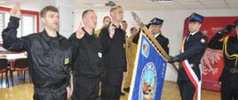 Świetlica komendy powiatowej. Przy pochylonym sztandarze trzech strażaków w ubraniach koszarowych koloru czarnego składa ślubowanie. Strażacy występują bez nakrycia głowy. Mają podniesione prawe ręce przedramieniem pionowo do góry, dłonie na wysokości oczu zwrócone w przód, z palcami ułozonymi tak jak do salutowania. Dowódca pocztu i asystujący salutują.