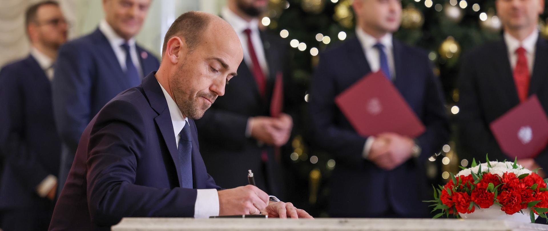 Minister aktywów państwowych Borys Budka podpisuje dokument. W tle członkowie Rady Ministrów. 