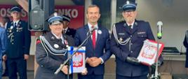 Mężczyzna w garniturze stoi po środku po jego lewej stronie stoi kobieta w mundurze strażackim i w dłoniach trzyma herb Polski, a po jego prawej stronie stronie stoi mężczyzna w mundurze strażackim i też trzyma w dłoniach herb Polski. 