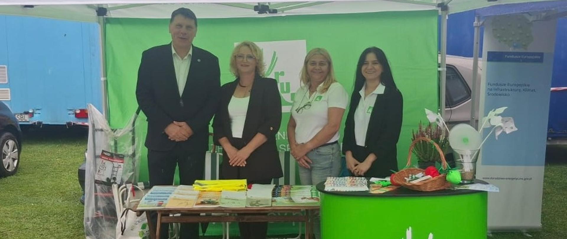 Stoisko informacyjne KRUS Placówki Terenowej w Miechowie pod zielono-białym namiotem Terenowej w Miechowie Edyta Włodarczyk-Gawron, Lesława Gawęcka, Wioleta Smolińska) jeden mężczyzna (dyrektor Małopolskiego Oddziału Regionalnego ARiMR Michał Chwastek) z przodu ustawiono zieloną ladę z logo KRUS i napisem pracuj bezpiecznie, na której znajdują się upominki dla dzieci.