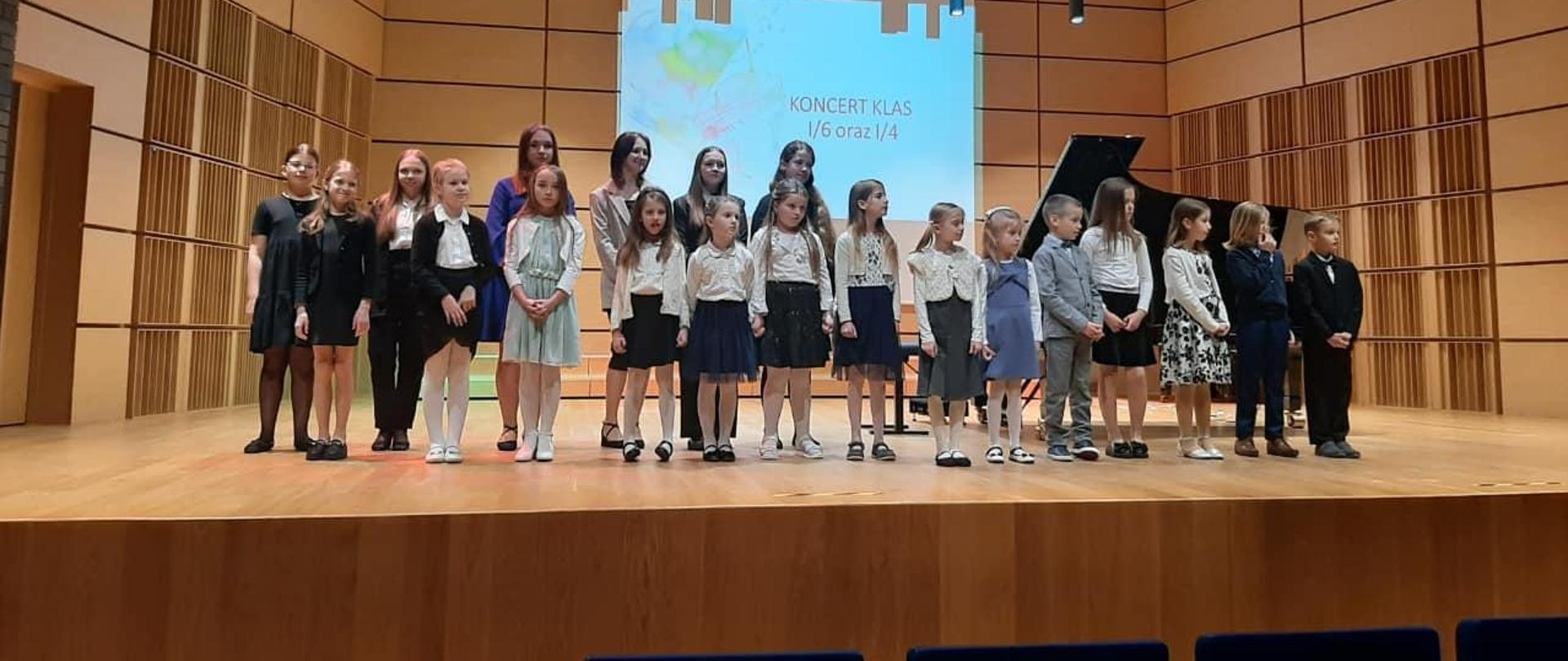 Uczniowie stojący w dwóch rzędach na scenie sali koncertowej. W tle wyświetlany napis: koncert I/6 oraz I/4 na projektorze. Z sufitu zwisają lampy.
