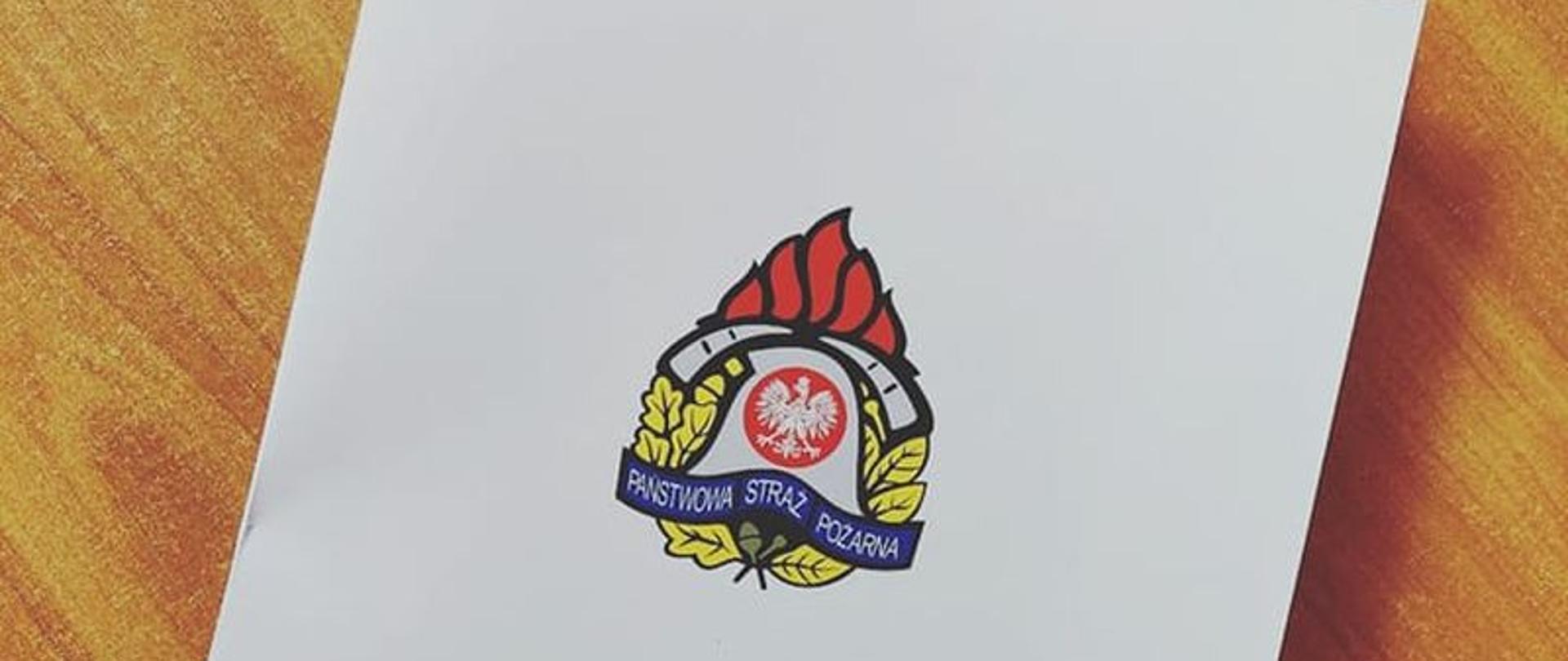 Zdjęcie przedstawia białą teczkę z logiem Państwowej Straży Pożarnej i podpisem Komenda Powiatowa Państwowej Straży Pożarnej w Białogardzie.Teczka leży na biurku.