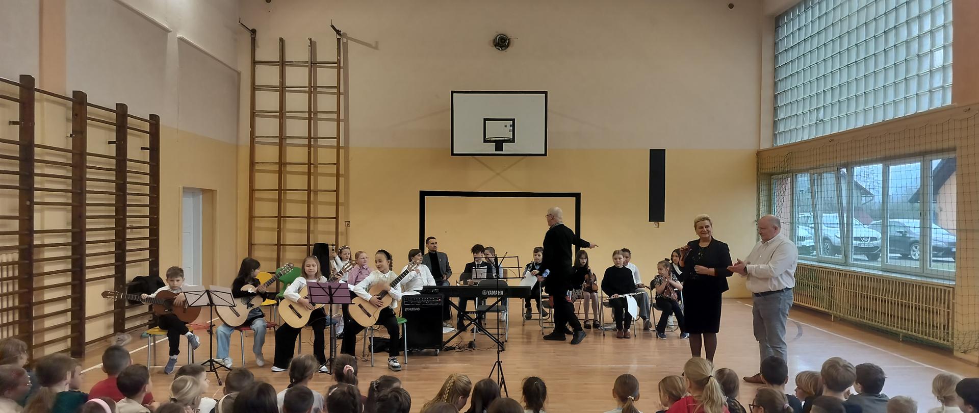 Uczniowie szkoły muzycznej siedzą w półkolu na środku sali, prezentując swoje umiejętności. Na pierwszym planie widać grupę gitarzystów, a w głębi m.in. pianistę przy instrumentach klawiszowych oraz innych instrumentalistów. Po prawej stronie stoją dorośli – prawdopodobnie dyrekcja lub nauczyciele obu placówek – którzy prowadzą spotkanie i dziękują wykonawcom. W centrum, tyłem do obiektywu, widoczny jest dyrygent lub opiekun artystyczny grupy. Na podłodze, przodem do młodych artystów, siedzi liczna grupa uczniów szkoły goszczącej, uważnie obserwując występ. Typowa sceneria szkolna – drabinki gimnastyczne, kosz do koszykówki oraz duże okna, przez które wpada dzienne światło.