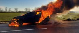 Pożar samochodu. Droga wojewódzka nr 434 w okolicach miejscowości Roszkówko. Na tle pola i drzew pali się ciemny samochód osobowy stojący na jezdni. Płomienie wydobywają się z przodu auta oraz przez tylne drzwi. Towarzyszy temu czarny dym.