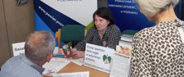 Starszy mężczyzna siedzący na krześle i stojąca obok niego starsza kobieta o siwych włosach, rozmawiają z kobietą siedzącą po drugiej stronie stołu. Kobieta o ciemnych włosach, trzyma w ręku smokerlyzer (urządzenia służącego do pomiaru tlenku węgla w wydychanym powietrzu). Za rozmawiającym, w tle, widoczne dwa banery, jeden z logo Państwowej Inspekcji Sanitarnej, drugi z niebieskim napisem: „Powiat Poznański” na białym tle i grafiką logo powiatu poznańskiego.
