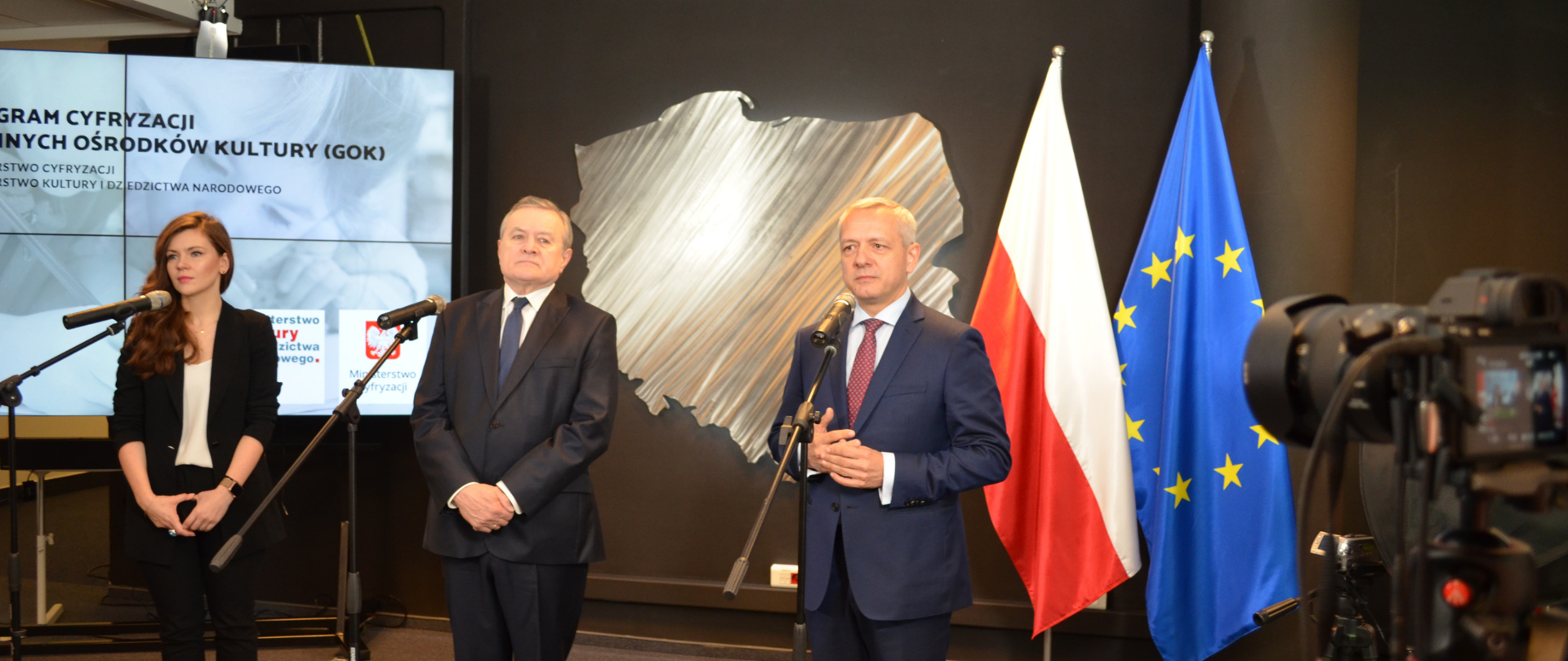 Konferencja prasowa. Przy mikrofonach stoją od lewej: Wanda Buk, Piotr Gliński , Marek Zagórski. W tle ekran z prezentacją, tablica w kształcie Polski, flagi polska i unijna. Po prawej stronie aparat fotograficzny nagrywający przebieg wydarzenia. 