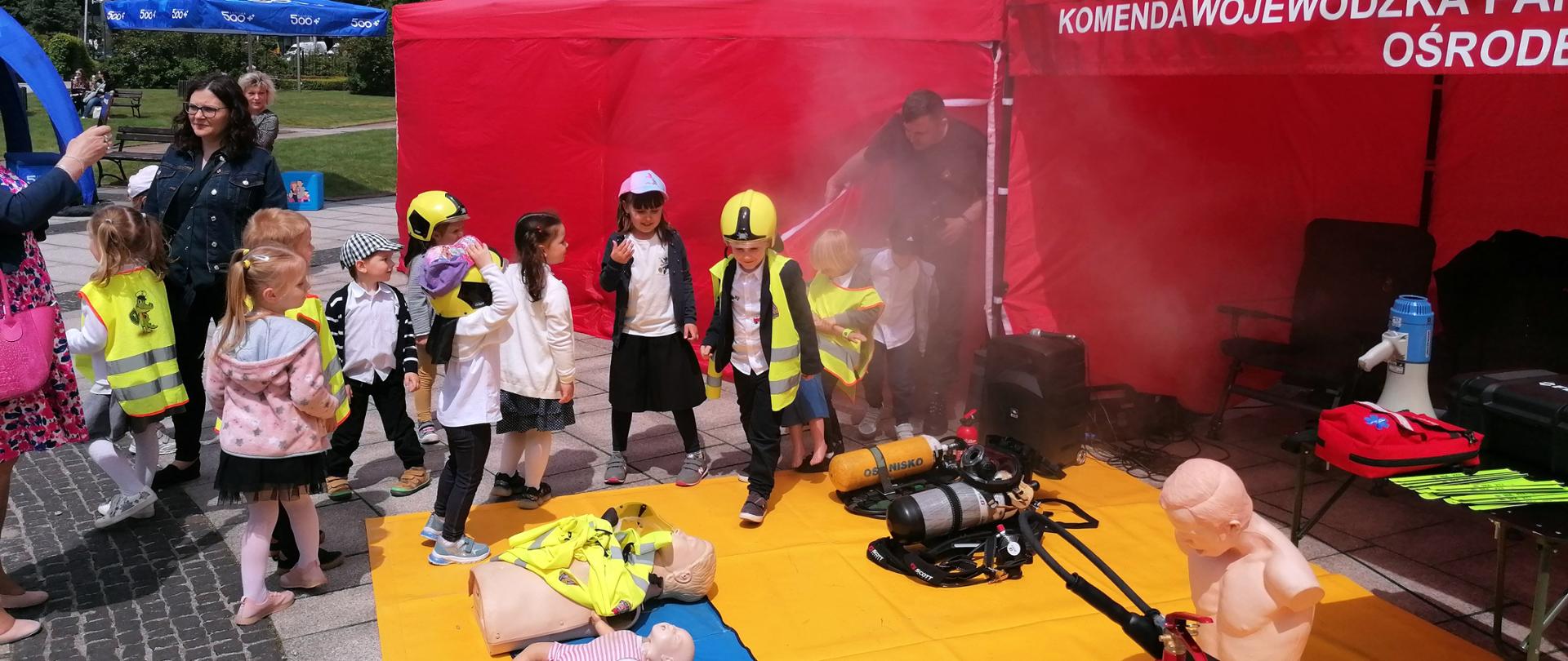 Na zdjęciu grupa dzieci zwiedzające stoisko Państwowej Straży Pożarnej na pikniku rodzinnym. Na stoisku wyeksponowane fantomy do prowadzenia szkolenia z pierwszej pomocy oraz aparaty powietrzne
