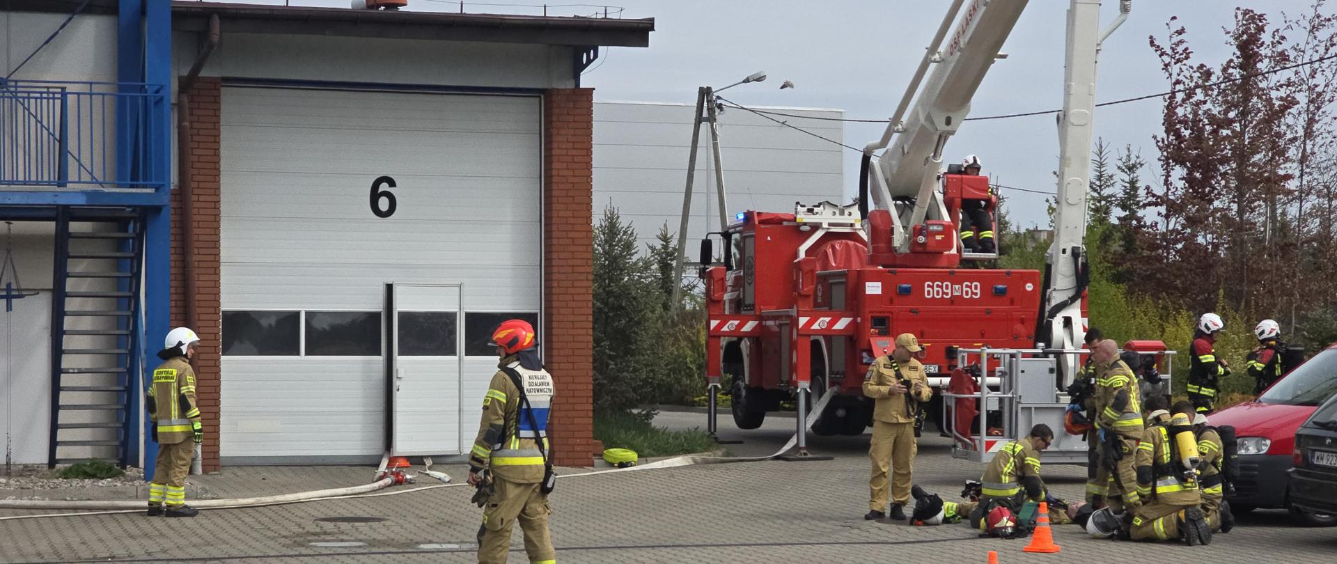 strażacy podczas ćiwczeń na placu przed JRG, w tle samochody osobowe, słupki ostrzegawcze i rozstawiony podnośnik